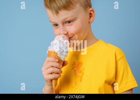 Positiver Junge, der Eis auf blauem Hintergrund leckt. Dreckiger Eisfleck auf gelber Kleidung. Daily Life Fleck und Reinigungskonzept Stockfoto