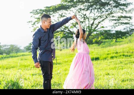 lateinischer Vater hält die Hand seiner Tochter, während sie tanzt, auf einem Grasfeld stehend und lächelnd. Stockfoto