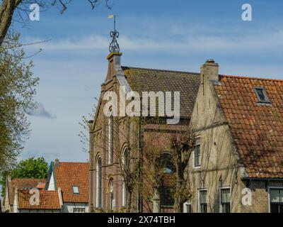 Reihe von alten Häusern mit roten Ziegeldächern, umgeben von Bäumen und einem klaren blauen Himmel, historische Häuser in einer kleinen Straße mit grünen Bäumen, Hollum Stockfoto