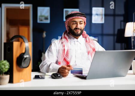 Arabischer Geschäftsmann im Heimbüro, der effizient auf seinem Laptop surft und tippt. Er arbeitet mit Online-Recherchen, Einkaufen und Zahlungen mit Kredit- und Debitkarten. Stockfoto