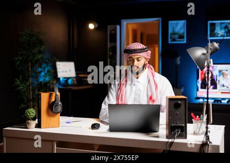 An einem modernen Arbeitsplatz tastet ein selbstbewusster arabischer Geschäftsmann auf seinem Laptop. Muslim Guy surft, redet per E-Mail und nutzt WLAN-Konnektivität in einer ruhigen und professionellen Geschäftsumgebung. Stockfoto