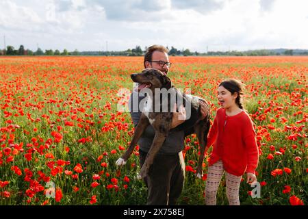 Porträt von glücklichem Vater und Tochter, die ihren Hund auf dem Mohnfeld umarmen Stockfoto