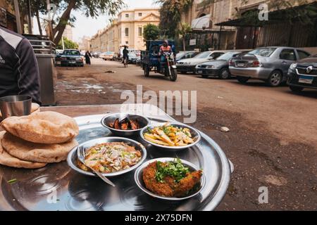 Ägyptisches Frühstück am Straßenrand im Zentrum von Kairo mit Falafel, Medames, Pommes, Auberginen und Baladi-Brot, serviert auf einem Metalltablett in der Nähe geparkter Autos Stockfoto