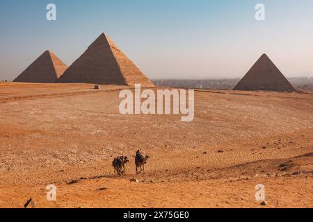 Kamelreiter überqueren die Wüste vor den drei Pyramiden von Gizeh – Khufu, hafre und Menkaure – unter klarem Himmel mit Kairos Dunst im Hintergrund Stockfoto