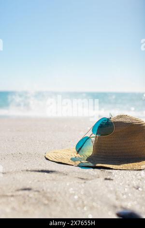 Sonnenhut und Sonnenbrille am Sandstrand; Wellen krachen vor blauem Hintergrund Stockfoto