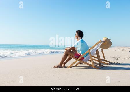 Am Strand sitzt ein junger, birassischer Mann auf einem Liegestuhl und blickt aufs Meer Stockfoto
