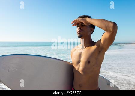 Am Strand, junger, birassischer Mann mit Surfbrett, der in die Ferne blickt Stockfoto