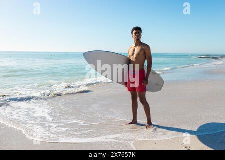 Am Strand, junger, birassischer Mann mit Surfbrett, stehend Stockfoto