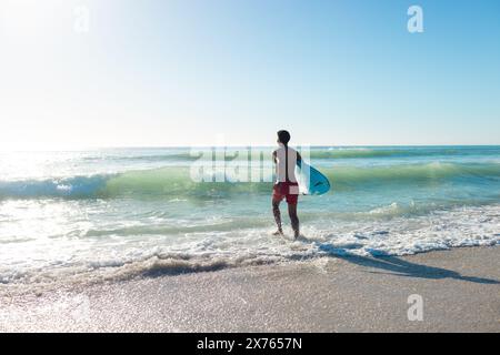 Am Strand, junger, birassischer Mann mit Surfbrett, der ins Meer geht Stockfoto