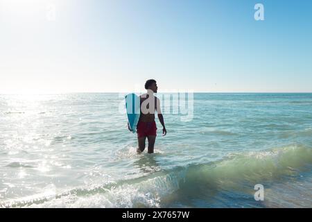 Am Strand spaziert ein birassischer junger Mann mit Surfbrett im Meer. Er trägt rote Shorts und blickt auf die Wellen, mit lockigen schwarzen Haaren, genießt den sonnigen Tag. Stockfoto