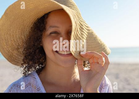 Am Strand, zweirassige junge Frau, die einen breitkrempigen Hut hält, lächelnd Stockfoto