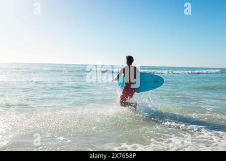 Am Strand läuft ein junger, birassischer Mann mit Surfbrett ins Meer Stockfoto