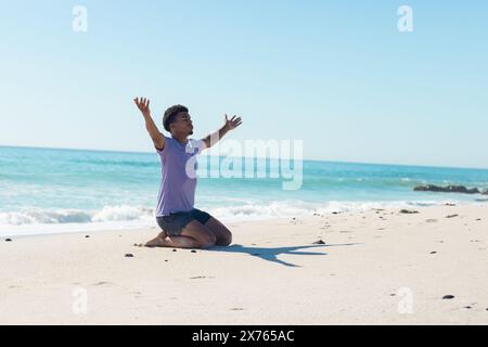 Am Strand sitzt ein junger, birassischer Mann und hebt die Hände hoch Stockfoto