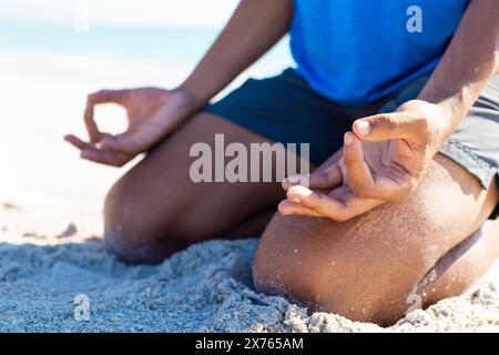 Am Strand sitzt ein junger, birassischer Mann, der in blauem Hemd meditiert Stockfoto