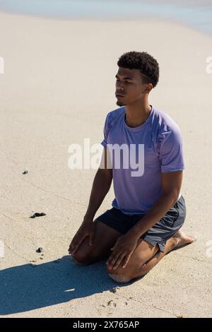 Am Strand sitzt ein junger, birassischer Mann auf Sand und blickt nachdenklich weg Stockfoto