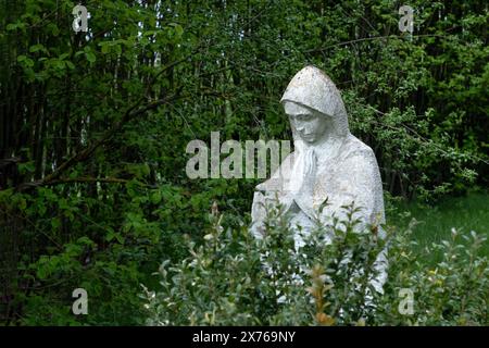 Eine beschädigte, alte Statue der Mutter Gottes, verlassen in der Friedhofskippe, verwischte Bäume mit grünen Blättern im Hintergrund. Stockfoto