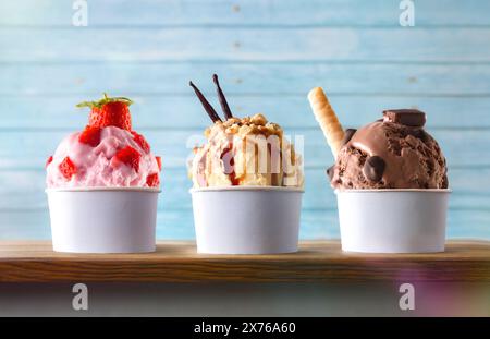 Drei Gläser Eis mit Erdbeeren-, Vanille- und Schokoladenkugeln auf einem Holztisch mit blauem Lattenhintergrund. Vorderansicht. Stockfoto