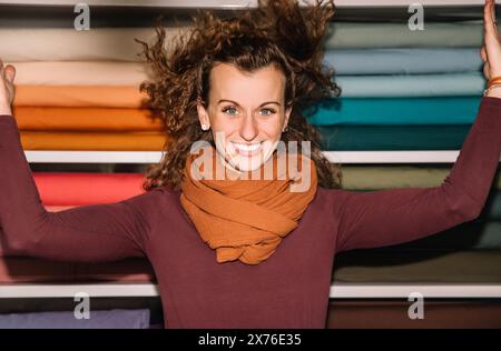 Lockige junge Frau, die breit lächelt, die Haare fliegen, in einem bunten Stoffgeschäft Stockfoto