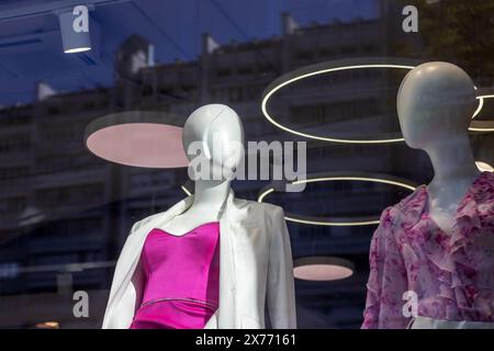 Zwei Schaufensterpuppen in einem Ladenfenster werden in lustigen pinken Kleidern und stilvollen Brillen präsentiert. Das Fashion-Design-Event ist mit einer Maskenhalterung in Magenta ausgestattet Stockfoto