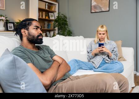 Ein junges Paar sitzt zu Hause auf dem Sofa, eine Frau ignoriert den Mann, schreibt SMS auf dem Smartphone, ein Mann blickt in die Ferne mit tiefen Gedanken. Relationshi Stockfoto