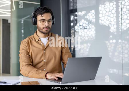 Ein fokussierter Mann mit Kopfhörern und Brillen, der an einem Laptop in einer modernen Büroumgebung arbeitet. Er konzentriert sich auf seine Aufgabe mit einem Notizblock und Tablet auf dem Tisch. Stockfoto