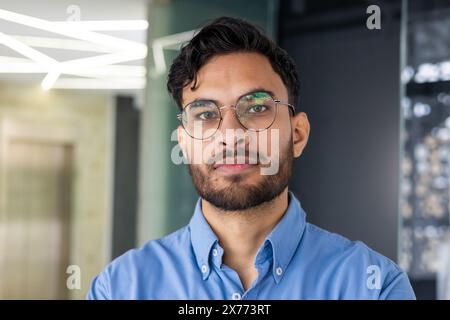 Selbstbewusster junger Mann mit Brille und Bart in einem blauen Hemd, der in einer modernen Büroumgebung steht. Stockfoto