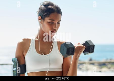 Indisches Mädchen, Ohrhörer und Hantel für Fitness im Freien, Bizeps und Training für Muskelwachstum. Weibliche Person, selbstbewusst und Musik für das Gewicht Stockfoto