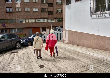 Zwei Frauen mit nicht erkennbarem Rücken gehen die Straße hinunter und unterhalten sich, eine in einem roten Trainingsanzug und die andere in einem weißen; beide haben graue Haare Stockfoto