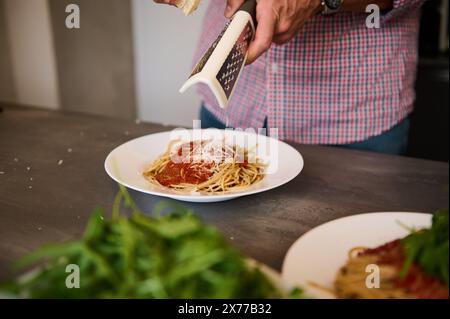 Nahaufnahme männlicher Hände, die Käse auf frisch gekochten italienischen Spaghetti mit Tomatensauce reiben und ein Familienessen in der Küche zubereiten. Italienische Küche. Fr Stockfoto