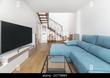Wohnzimmer mit einem großen Sofa mit blauem Stoff, einigen lackierten Holzstühlen, einem großen Fernseher und einer Treppe, die zu einem Dachgeschoss führt Stockfoto