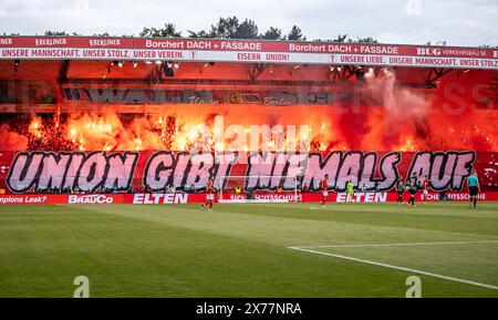 Berlin, Deutschland. Mai 2024. Fußball: Bundesliga, 1. FC Union Berlin - SC Freiburg, Spieltag 34, an der Alten Försterei. Die Fans der Union Berlin entzünden ein bengalisches Feuer auf der Waldseite hinter dem Schild „Union gibt nie auf!“ Hinweis: Andreas Gora/dpa – WICHTIGER HINWEIS: gemäß den Vorschriften der DFL Deutscher Fußball-Liga und des DFB Deutscher Fußball-Bundes ist es verboten, im Stadion und/oder des Spiels aufgenommene Fotografien in Form von sequenziellen Bildern und/oder videoähnlichen Fotoserien zu verwenden oder zu nutzen./dpa/Alamy Live News Stockfoto