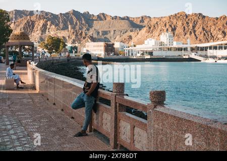 Ein Mann lehnt sich an eine Mauer an der Corniche im Sultan Qaboos Port, Maskat, Oman, bei Sonnenuntergang Stockfoto