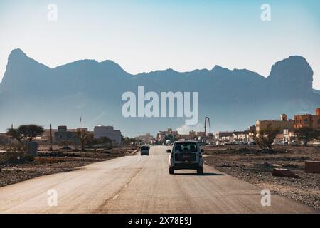 Fahrzeug fährt auf einer asphaltierten Straße durch eine ländliche Siedlung mit dramatischen Al Hajar Berggipfeln im Hintergrund, im Norden des Oman Stockfoto