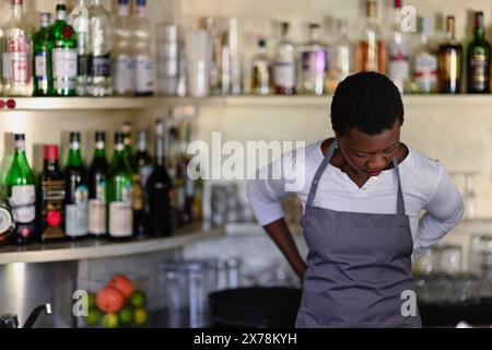Konzentrierter Barkeeper mit Schürze steht vor einer Vielzahl von Flaschen in einer voll ausgestatteten Bar Stockfoto