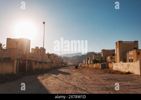 Sonnendurchflutete Wohngegend in der Nähe von Mughsail Beach, Oman, mit traditionellen Häusern, staubigen Straßen und Bergen in der Ferne Stockfoto