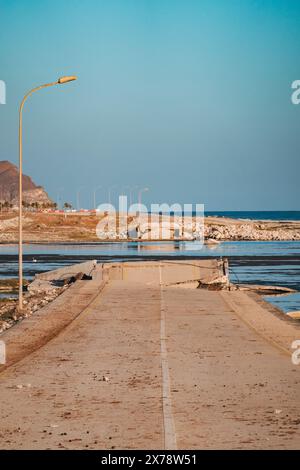 Eingestürzte Brücke am Al Mughsail Beach in Oman, mit abgebrochenen Straßenabschnitten, die vor einer Bergkulisse in seichtes Wasser führen. Stockfoto