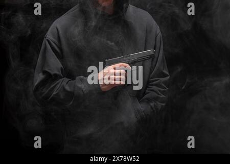 Junger Mann in einem schwarzen Hoodie mit einer Pistole auf einem rauchigen schwarzen Hintergrund Stockfoto