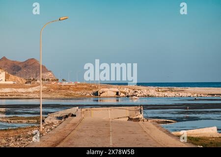 Eingestürzte Brücke am Al Mughsail Beach in Oman, mit abgebrochenen Straßenabschnitten, die vor einer Bergkulisse in seichtes Wasser führen. Stockfoto