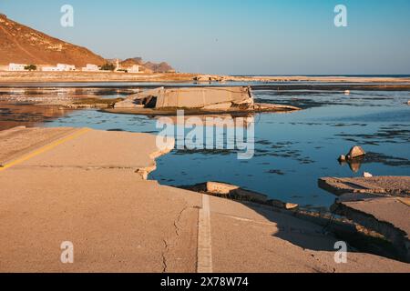 Eingestürzte Brücke am Al Mughsail Beach in Oman, mit abgebrochenen Straßenabschnitten, die vor einer Bergkulisse in seichtes Wasser führen. Stockfoto