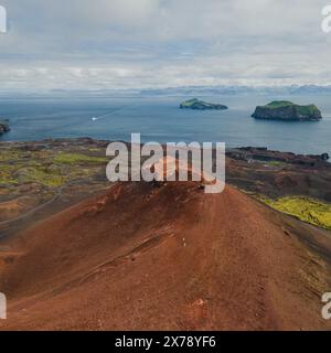 Aus der Vogelperspektive der vulkanischen Landschaft in Vestmannaeyjar (Westman Islands), Island, mit Blick auf das zerklüftete Gelände, die roten vulkanischen Hügel und den entfernten isl Stockfoto
