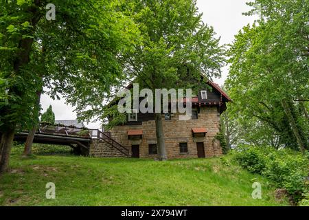 Ein Steinhaus mit einer hölzernen Veranda liegt in einem üppigen grünen Feld. Das Haus ist von Bäumen umgeben und hat eine Brücke, die hinauf führt. Die Szene ist Peacefu Stockfoto