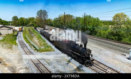 Ein luftiger, leistungsstarker Oldtimer-Dampfzug fährt durch einen Industriebahnhof und emittiert Dampf, während er durch Lagerhäuser und neben anderen Eisenbahnwaggons und vorbeifährt Stockfoto