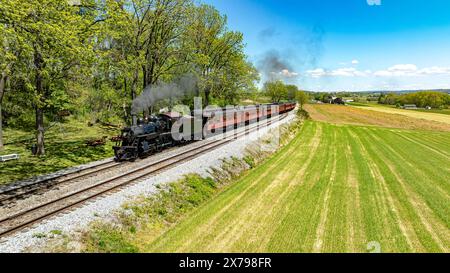 Ein bezaubernder Blick aus der Vogelperspektive auf eine historische Dampfeisenbahn, die eine Rauchwolke ausstrahlt, während sie durch eine üppige Landschaft fährt, flankiert von einem lebhaften grünen Feld Stockfoto