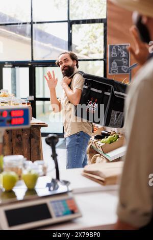 Kaukasischer Kurier verlässt einen umweltfreundlichen 24-Stunden-Laden, um lokal angebaute Produkte an die Kunden zu liefern. Junger Liefermann mit Thermotasche und Mitnahme aus nachhaltigem Supermarkt. Stockfoto