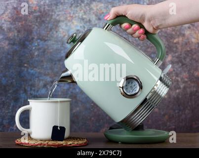 Eine Frau gießt heißes Wasser aus einem Wasserkocher in eine Tasse mit einem Einweg-Teebeutel. Kopierbereich. Stockfoto