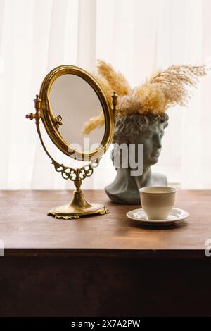 Eleganter Vintage-Spiegel und Büstenvase mit Pampas-Gras neben einer Kaffeetasse auf einem Holztisch Stockfoto