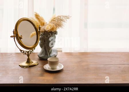 Eleganter Vintage-Spiegel und Büstenvase mit Pampas-Gras neben einer Kaffeetasse auf einem Holztisch Stockfoto