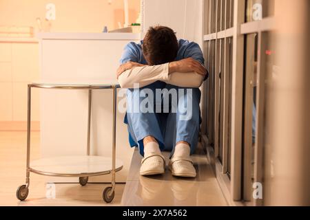 Gestresste männliche Krankenschwester, die abends im Krankenhaus auf der Fensterbank sitzt Stockfoto