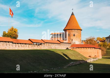 Die Burg Kaunas zeigt ihre markanten roten Türme, das lebhafte Herbstlaub und die litauische Flagge, die im späten Nachmittagslicht gegen einen klaren Himmel winkt Stockfoto