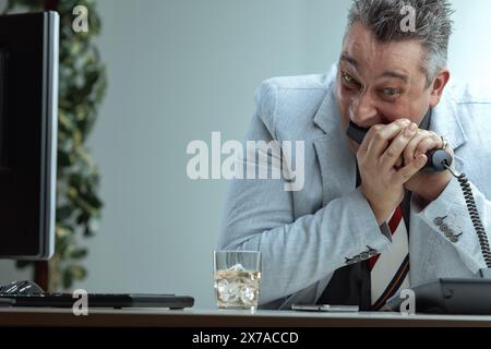 Mit leichtem Anzug und gestreifter Krawatte isst der Mann ein Telefon mit frustriertem Ausdruck. Ein Glas Whiskey auf seinem Schreibtisch zeigt den Stress von Managi Stockfoto